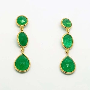 May include: A pair of gold earrings with three green gemstones each. The top gemstone is round, the middle is oval, and the bottom is teardrop shaped.