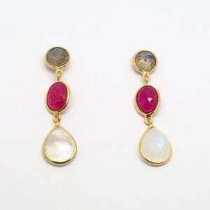 May include: A pair of gold drop earrings. Each earring has three gemstones: a small, round, grey stone at the top, a large, oval, red stone in the middle, and a pear-shaped, white stone at the bottom. The earrings are set against a white background.