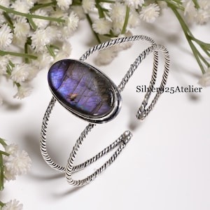 Può includere: Un bracciale a fascia in argento con una grande pietra preziosa di labradorite di forma ovale con iridescenza viola e blu. Il cinturino è composto da due fili d'argento intrecciati che si uniscono alla montatura. Il testo Silver25Atelier è visibile.