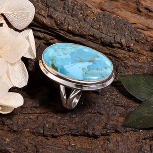 Peut inclure: Une bague en argent ornée d'une grande pierre turquoise de forme ovale avec des motifs marbrés bleu clair et bleu foncé. La pierre est sertie dans une monture en argent lisse et poli, et l'anneau est composé de deux fins fils d'argent courbés.
