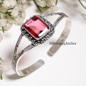 Pode incluir: Uma pulseira de prata com uma grande pedra preciosa rosa retangular e facetada, inserida numa moldura decorativa e ornamentada com padrões intrincados de volutas ao longo da banda. O texto Silver925Atelier está impresso na banda de metal.