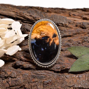 May include: An oval ring featuring a polished gemstone with a swirling pattern of deep black and warm amber tones. The stone is set in a silver-toned metal frame with a twisted rope-style border, creating a distinct contrast between the dark and light colors.