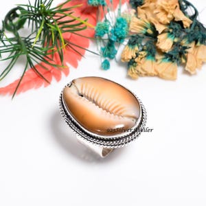 Può includere: Un anello con una conchiglia di ciprea ovale e lucida, nei toni, nei toni del marrone e crema, incastonata in una montatura in argento sterling testurizzato con un bordo a corda intrecciata. L'anello in argento reca la scritta 925SilverAtelier.