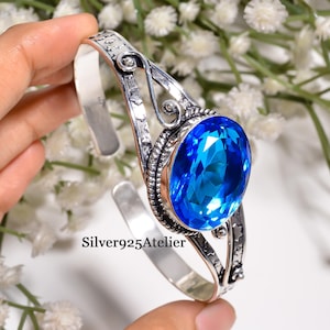 Puede incluir: Un brazalete de plata esterlina con una gran piedra preciosa azul vibrante de corte ovalado, engastada en un marco decorativo con intrincados adornos de volutas y recortes en forma de estrella a lo largo de la banda de metal pulido.