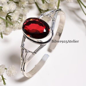 Può includere: Un bracciale a fascia in argento sterling con una grande pietra granato rosso scuro dal taglio ovale, incastonata in una montatura in metallo lucido. Il bracciale ha un design diviso con piccole sfere decorative in metallo. Il testo Silver925Atelier è visibile.