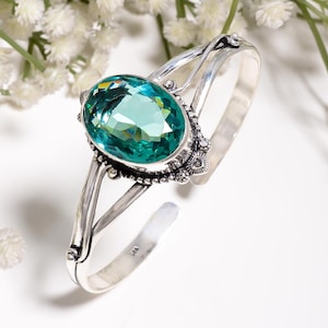 Op de afbeelding: Een zilveren armband met een grote, ovaal geslepen, gefacetteerde blauwgroene edelsteen in een decoratieve, met kralen versierde metalen zetting. De band bestaat uit twee gespleten zilveren armen die aan de zijkanten samenkomen.