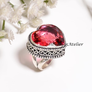 Puede incluir: Un anillo de plata de ley con una gran piedra preciosa rosa tallada en forma de corazón, engastada en un bisel decorativo con volutas intrincadas y bordes de cuentas. La banda está dividida en dos. El texto Silver925Atelier es visible.