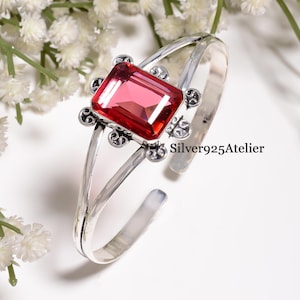 Op de afbeelding: Een armband van sterling zilver met een grote, rechthoekige, gefacetteerde rode edelsteen in het midden. De zetting bevat decoratieve zilveren krulmotieven rondom de steen. De tekst Silver925Atelier staat aan de rechterkant van de band.