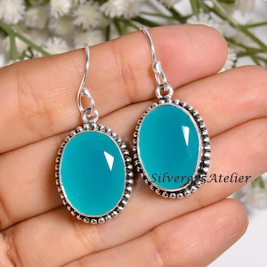 Peut inclure: Une paire de boucles d'oreilles pendantes en argent sterling avec des pierres précieuses ovales en calcédoine bleu sarcelle à facettes. Chaque pierre est sertie dans un cadre en argent perlé avec un crochet. Le texte SilverosAtelier est visible.