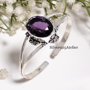 Peut inclure: Un bracelet manchette en argent sterling orné d'une grande pierre précieuse améthyste violette de taille ovale au centre. Le métal présente des détails complexes en volutes et floraux autour de la pierre. La bande intérieure porte le poinçon 925.