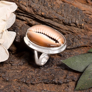 May include: A ring featuring a polished, oval-shaped brown and cream cowrie shell set into a smooth, silver-toned metal bezel. The band is constructed from a single piece of silver-toned metal that splits into two distinct sections at the base.