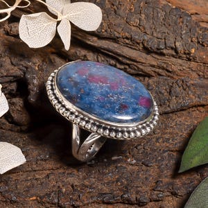 Peut inclure: Une bague en argent ornée d'une grande pierre cabochon ovale, présentant une base bleu profond avec des inclusions magenta dispersées. La pierre est sertie dans une monture en argent poli avec un bord perlé, sur un anneau argenté divisé.