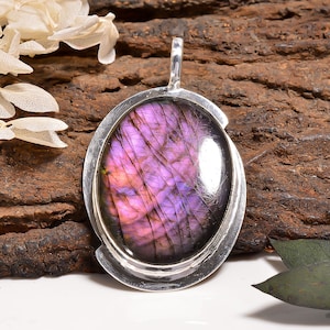 Può includere: Un ciondolo di forma ovale con una pietra preziosa di labradorite lucida con vivaci inclusioni viola e scure, incastonato in una cornice di metallo color argento liscia e riflettente con un gancio verticale sulla parte superiore.