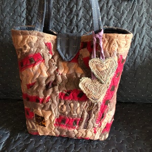 Może przedstawiać: Torba typu tote wykonana z patchworku z niebieskim uchwytem z dżinsu i dwoma zawieszkami w kształcie serca. Torba jest wykonana z różnych tkanin w odcieniach brązu, różu i czerwieni.