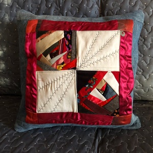 May include: A square pillow with a red satin border and a patchwork design. The patchwork is made of various fabrics in shades of red, white, black, and gray. The pillow is quilted with white thread.