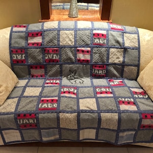 Könnte beinhalten: Eine graue und blaue Patchwork-Decke mit einem weißen gestickten Hasen in der Mitte. Die Decke besteht aus verschiedenen Strickstoffen und hat auf einigen Quadraten rot-weiße Streifen. Die Decke liegt auf einem weißen Sofa mit zwei Kissen.