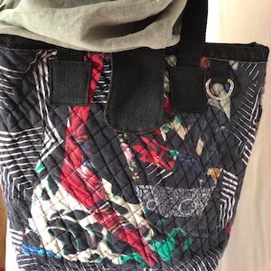 Könnte beinhalten: Eine schwarze gesteppte Tragetasche mit einem bunten geometrischen Muster. Die Tasche hat einen schwarzen Trageriemen und einen silbernen Ring oben.