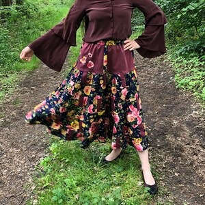 Könnte beinhalten: Eine bordeauxfarbene Bluse mit ausgestellten Ärmeln und Knopfleiste wird mit einem langen, fließenden Rock kombiniert. Der Rock hat ein Patchwork-Design mit floralen Mustern in Rosa, Gelb und Marineblau, was eine Bohème-Ästhetik erzeugt.