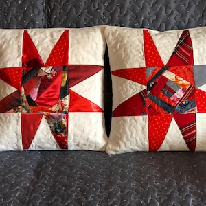 Op de afbeelding: Twee vierkante decoratieve kussens met een sterrenpatroon. De sterren zijn gemaakt van rode stof met verschillende patronen, waaronder stippen en bloemenprints, tegen een witte, gewatteerde achtergrond. De kussens zijn een gezellig woonaccessoire.