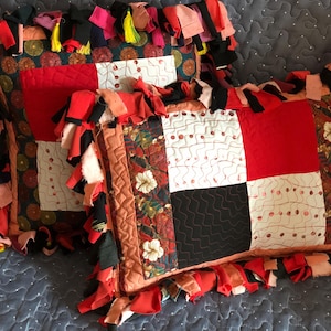 Op de afbeelding: Twee decoratieve sierkussens met patchwork-ontwerpen. De kussens hebben rode, zwarte, witte en bedrukte stoffen vierkanten. De randen zijn afgewerkt met kleurrijke stoffen stroken. De kussens liggen op een blauw gewatteerd oppervlak.