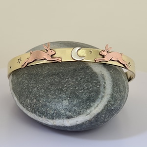 May include: A gold bangle bracelet featuring two copper rabbits and a silver crescent moon charm. The bracelet is decorated with small stars.