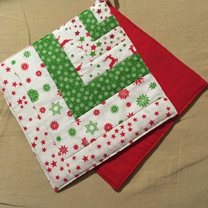 Könnte beinhalten: Ein gesteppter Weihnachts-Topflappen mit roter Rückseite und einer Schlaufe zum Aufhängen. Die Vorderseite hat ein Patchwork-Design mit grünem und weißem Stoff, verziert mit roten Sternen, grünen Sternen, Schneeflocken und Rentieren.