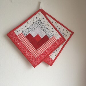 Könnte beinhalten: Zwei rote und weiße gequiltete Topflappen mit Patchwork-Muster. Die Topflappen hängen an einer weißen Wand.
