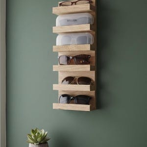 Puede incluir: Un organizador de pared vertical de madera clara con cinco estantes horizontales. Los estantes sostienen varios pares de gafas y dos estuches protectores blancos transl&uacute;cidos, uno de los cuales tiene la etiqueta con el texto BRILLEZA.