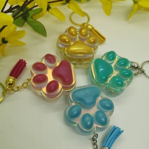 Puede incluir: Cuatro llaveros con forma de huella de pata iridiscente con detalles dorados, rosados, azul verdoso y azules. Cada llavero tiene un pequeño borla y un anillo de llave plateado.