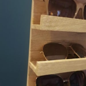 Puede incluir: Un expositor vertical de madera clara con m&uacute;ltiples estantes horizontales dise&ntilde;ados para guardar gafas. El expositor tiene un acabado de veta de madera natural y muestra varios pares de gafas de sol con lentes oscuras en los estantes.