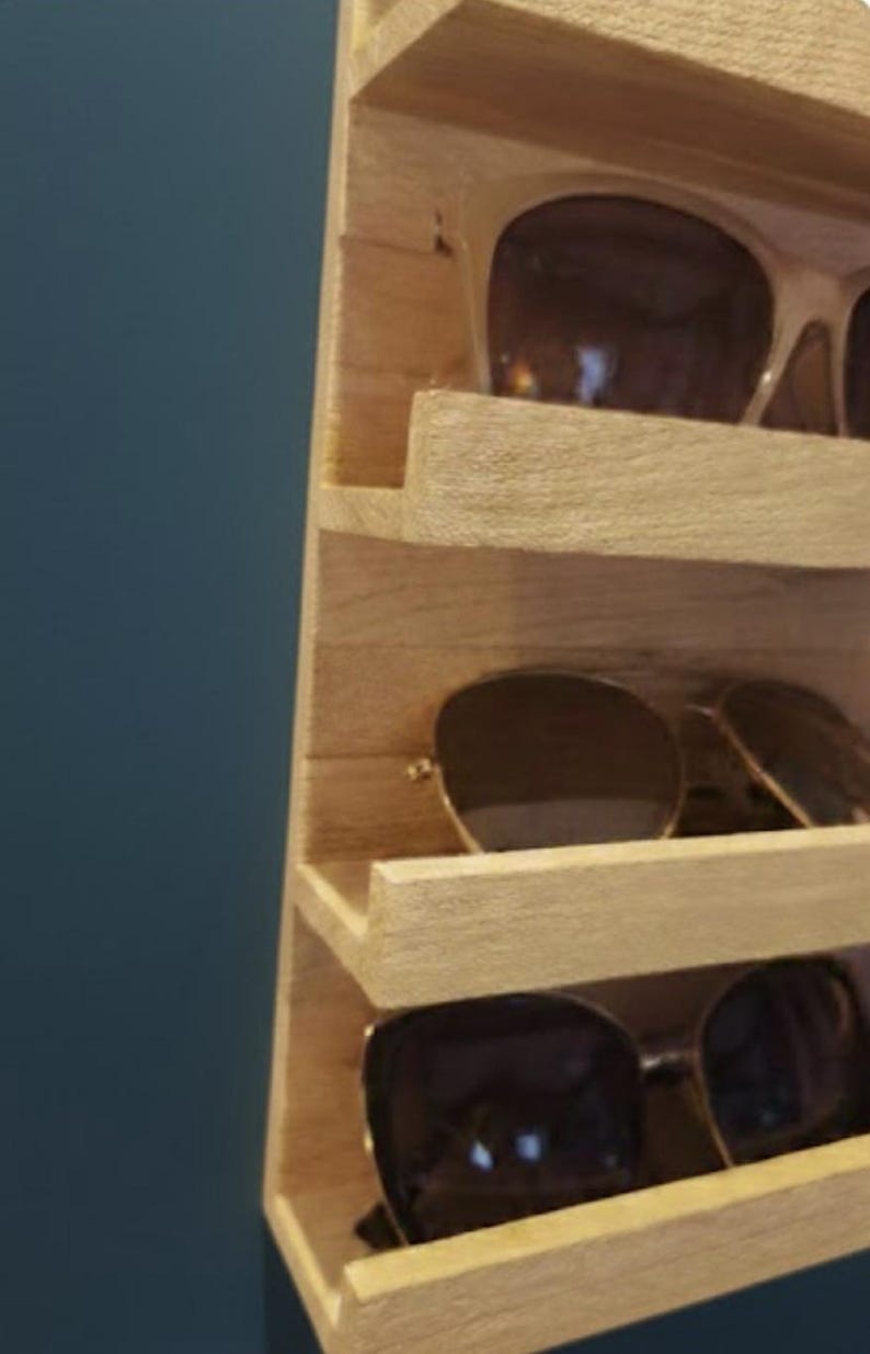 Puede incluir: Un expositor vertical de madera clara con m&uacute;ltiples estantes horizontales dise&ntilde;ados para guardar gafas. El expositor tiene un acabado de veta de madera natural y muestra varios pares de gafas de sol con lentes oscuras en los estantes.