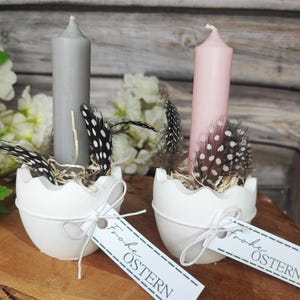 Könnte beinhalten: Zwei Osterkerzenhalter mit hohen Kerzen und Federn. Eine Kerze ist grau, die andere rosa. Die Halter sind weiß, eiförmig und haben ein Etikett mit "Frohe OSTERN".