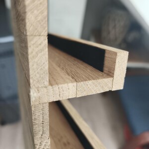 Puede incluir: Una estanter&iacute;a de madera de color claro con dos niveles horizontales y vetas visibles. Cada nivel de estante incluye una tira negra estrecha y empotrada a lo largo del borde posterior, que contrasta con el acabado de madera natural.