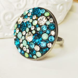 Könnte beinhalten: Silberner Ring mit einer großen, runden Oberfläche, die mit einem Mosaik aus blauen, türkisfarbenen und klaren Kristallen bedeckt ist. Die Kristalle variieren in Größe und Form und erzeugen eine glitzernde, strukturierte Oberfläche. Der Ring ist vor einem weichen, weißen Hintergrund platziert.