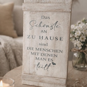 Puede incluir: Un letrero rectangular de madera con el texto alemán "Das Schönste an zu Hause sind die Menschen mit denen man es teilt" en letras grises oscuras. El letrero está pintado de blanco con un acabado envejecido, sobre una superficie de madera.