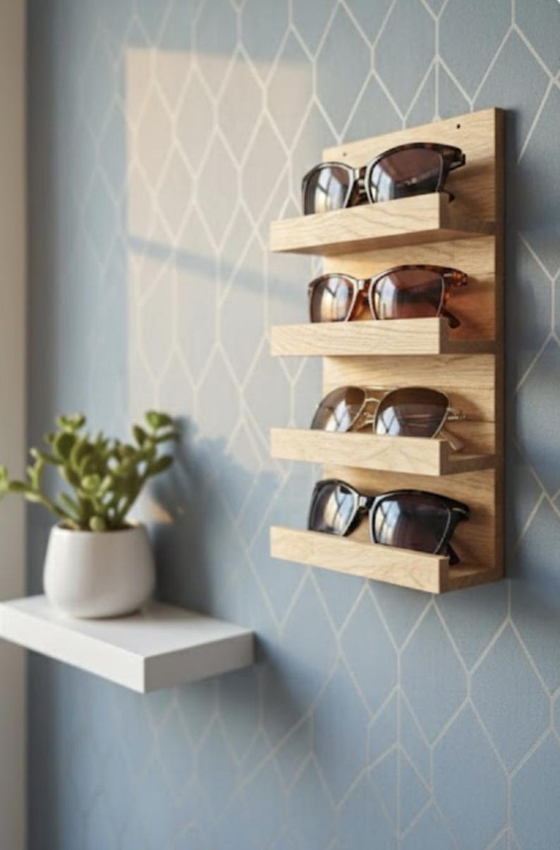 Puede incluir: Un estante de pared vertical de madera clara con cuatro repisas horizontales para almacenamiento. Cada repisa sostiene un par de gafas de sol con lentes y monturas oscuras. El estante est&aacute; montado en una pared con un patr&oacute;n geom&eacute;trico.