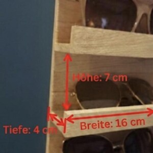 Puede incluir: Un estante de pared de madera clara con m&uacute;ltiples repisas horizontales para guardar gafas. Textos y flechas rojas indican dimensiones de 7 cm de altura, 16 cm de ancho y 4 cm de profundidad. El estante contiene varios pares de gafas de sol.