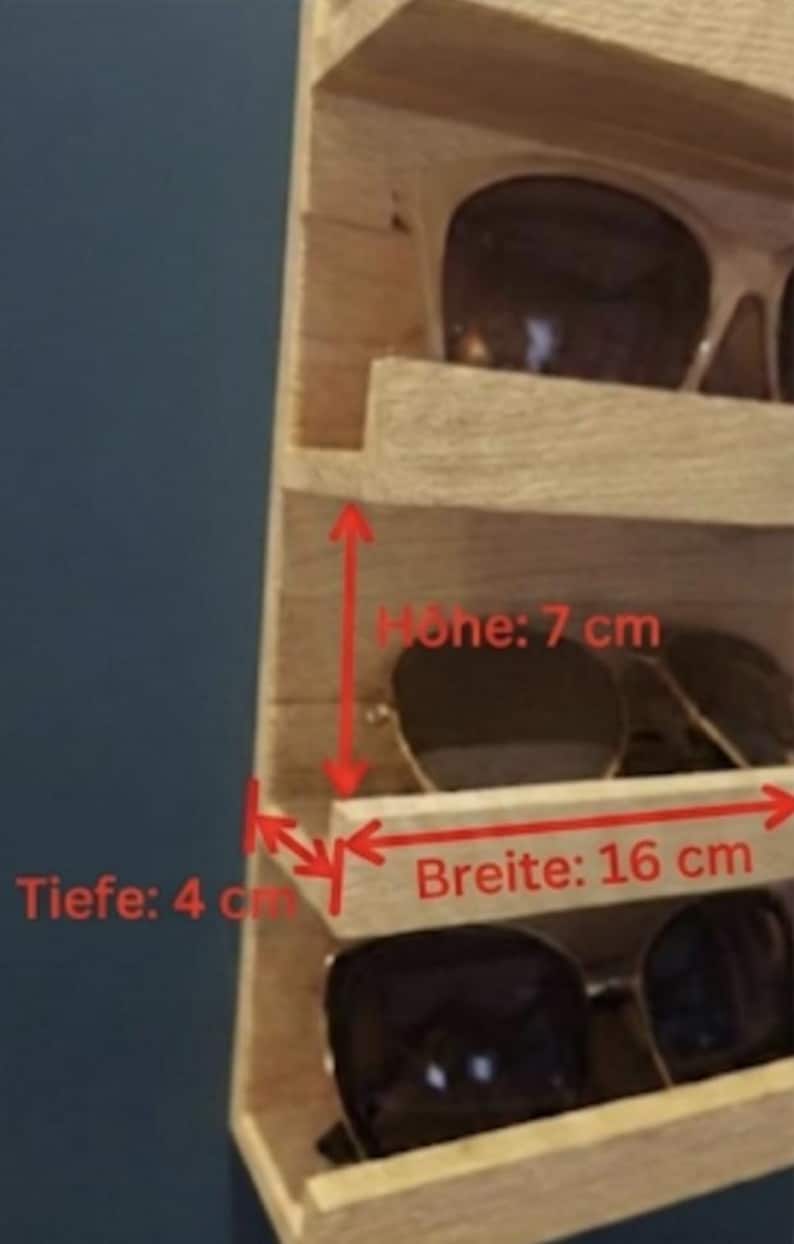 Puede incluir: Un estante de pared de madera clara con m&uacute;ltiples repisas horizontales para guardar gafas. Textos y flechas rojas indican dimensiones de 7 cm de altura, 16 cm de ancho y 4 cm de profundidad. El estante contiene varios pares de gafas de sol.
