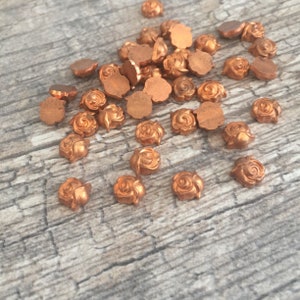 May include: A collection of small, copper-colored metal rose embellishments scattered on a wooden surface. The roses have detailed petals and a flat back. These decorative elements are ideal for crafting and jewelry making.