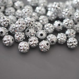 May include: A collection of silver-colored, faceted beads. The beads are spherical with a textured surface, reflecting light. The beads are scattered, creating a sparkling effect, suitable for jewelry making or craft projects.