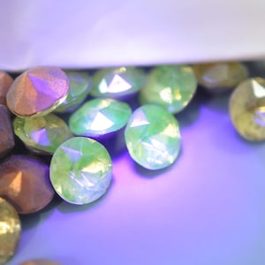 Puede incluir: Primer plano de gemas facetadas e iridiscentes en tonos verdes, dorados y bronce. Las gemas ovaladas parecen estar dispersas sobre una superficie p&uacute;rpura. La iluminaci&oacute;n crea reflejos y sombras, realzando el brillo.