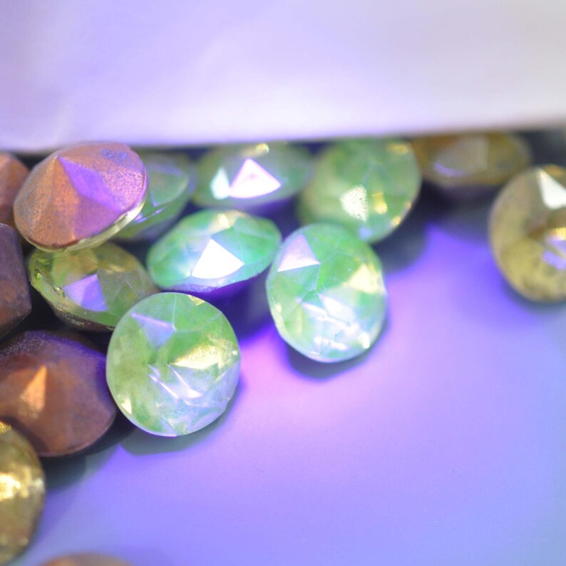 Puede incluir: Primer plano de gemas facetadas e iridiscentes en tonos verdes, dorados y bronce. Las gemas ovaladas parecen estar dispersas sobre una superficie p&uacute;rpura. La iluminaci&oacute;n crea reflejos y sombras, realzando el brillo.