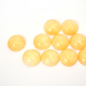 May include: A group of ten round, yellow, translucent cabochons with a swirl pattern. The cabochons are arranged in a semi-circle on a white surface.