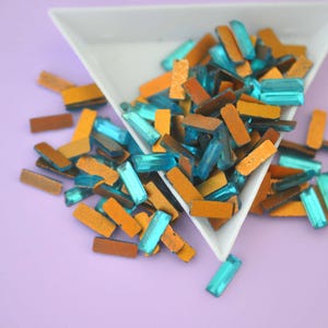 May include: A close-up of a white triangular dish filled with turquoise and gold rectangular glass rhinestones. The rhinestones are scattered on a purple surface.