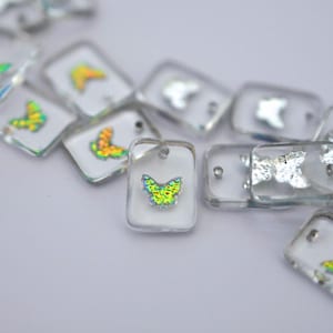 May include: Clear rectangular pendants with iridescent butterfly designs. The pendants have a small hole at the top for stringing. The butterflies have yellow, green, and blue coloring. The pendants are scattered on a white surface.