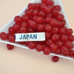 May include: A white triangular dish filled with numerous small, translucent red beads. A label reading "JAPAN" is visible. The beads are clustered together, creating a vibrant, eye-catching display.