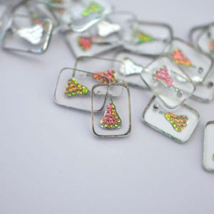 May include: A collection of clear, rectangular charms with iridescent, colorful embellishments. Each charm has a silver-colored frame and a small, decorative design in the center, creating a sparkling effect. The charms are scattered on a white surface.