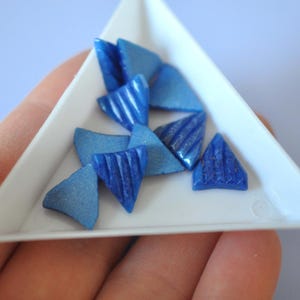 May include: A collection of blue triangle-shaped cabochons with a striated texture. The cabochons are arranged in a white triangular tray.