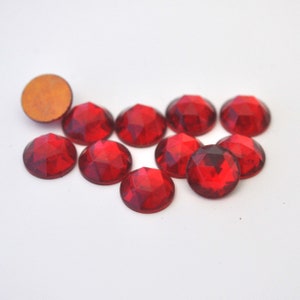 May include: A collection of ten faceted, round, red rhinestones. One rhinestone is turned over, revealing a tan backing. The rhinestones are arranged on a white surface, showcasing their vibrant color and geometric design.