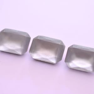 May include: Three rectangular, faceted, grey gemstones are arranged in a row against a light purple background. The gemstones have a subtle, translucent quality, with a slightly frosted appearance. The facets catch the light, creating subtle highlights.